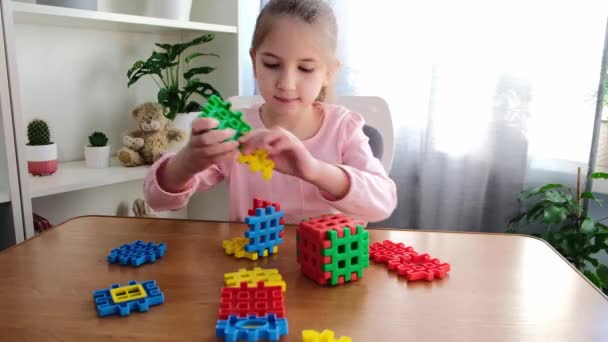 Mała dziewczynka w wieku 6-7 lat w różowej koszulce siedzi przy stole z misiaczkiem w domu, zbiera puzzle-konstruktor. — Wideo stockowe