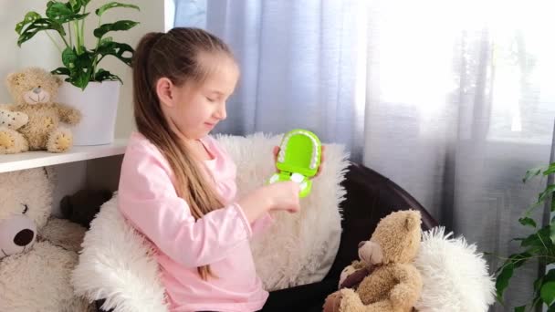 Маленькая девочка с длинными волосами, одетая в розовую футболку, играет дома на стуле с игрушками, показывающими, как правильно чистить зубы. Концепция здорового образа жизни — стоковое видео