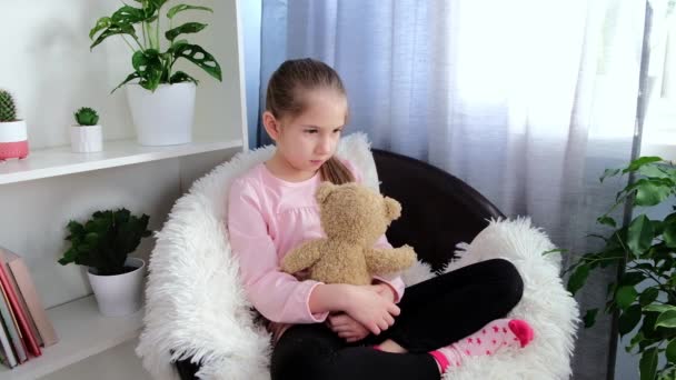 Сумна дівчина 6-7 років в рожевій футболці сидить на кріслі вдома обіймаючи плюшевого ведмедя — стокове відео