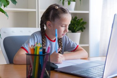 A 6-8-year-old girl in a gray T-shirt to study at the computer at home, studies, does homework, communicates on the Internet, writes in a notebook.
