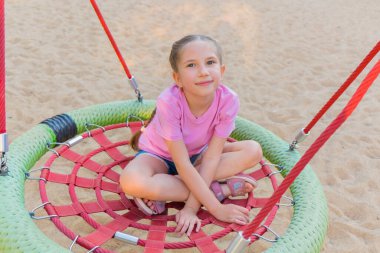 7-8 yaşlarında, uzun saçlı, pembe tişört ve şortlu, örümcek ağı yuvasında gülümseyen bir kız lunaparkta sallanıyor..