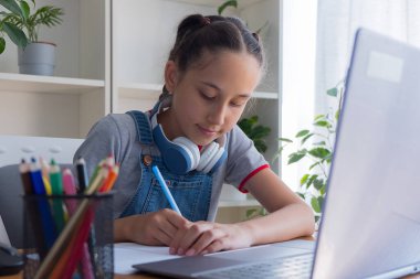 A 11-13-year-old girl in a gray T-shirt uses headphones to study at the computer at home, studies, does homework, communicates on the Internet, writes in a notebook.