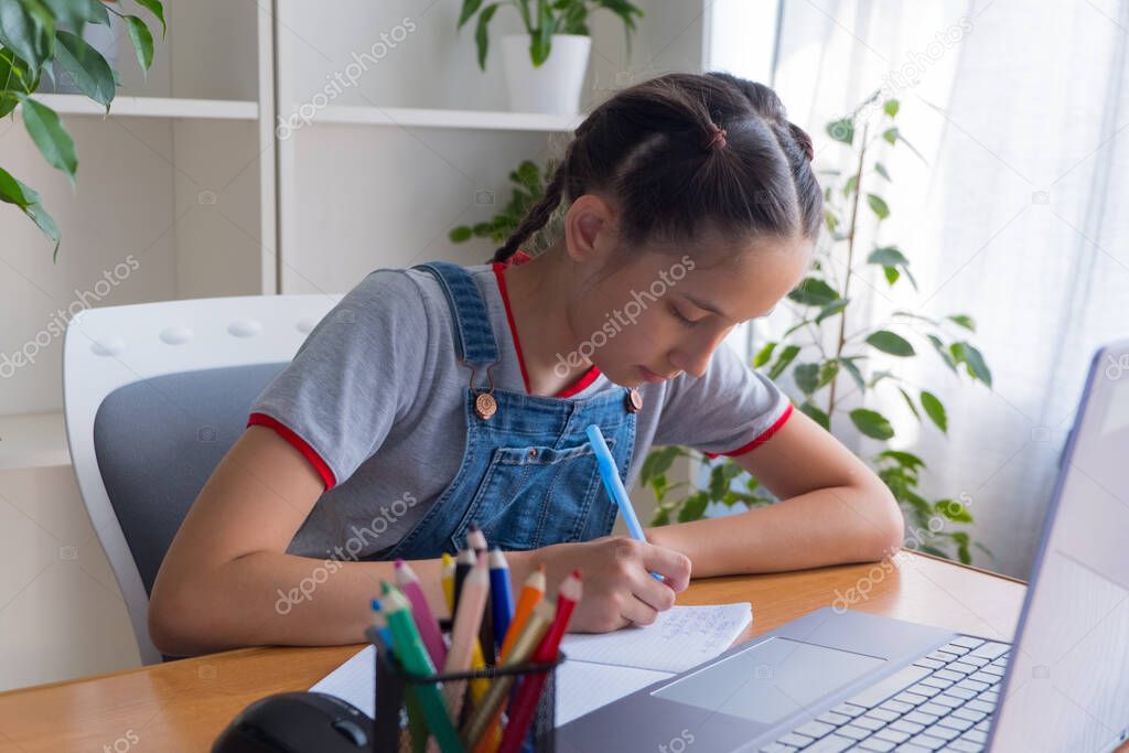 A brunette 11-13 year-old girl in a gray T-shirt to study at the computer at home, studies, does homework, communicates on the Internet, writes in a notebook.