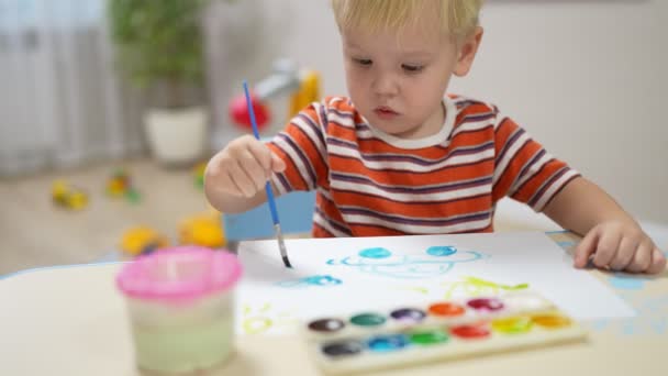 A little boy with blond hair sits at a table and draws a car with paints on paper — Stockvideo