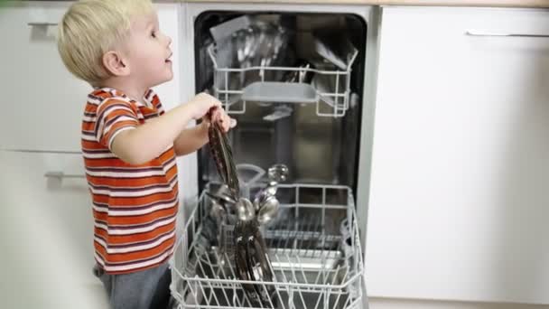 Niño pequeño con el pelo rubio se sienta ayudar a mamá a sacar los platos del lavavajillas — Vídeos de Stock