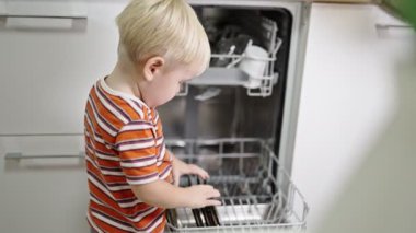 little boy with blond hair helps mom get the plates out of the dishwasher.