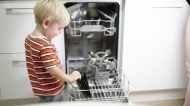 Sarı saçlı küçük çocuk annesinin bulaşık makinesinden bulaşıkları çıkarmasına yardım ediyor..