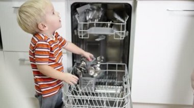 Sarı saçlı küçük çocuk annesinin bulaşık makinesinden bulaşıkları çıkarmasına yardım ediyor..