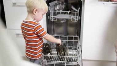 Sarı saçlı küçük çocuk annesinin bulaşık makinesinden bulaşıkları çıkarmasına yardım ediyor..