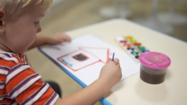 Niño pequeño con el pelo rubio se sienta en la mesa y pinta con pinturas en la casa de papel — Vídeos de Stock