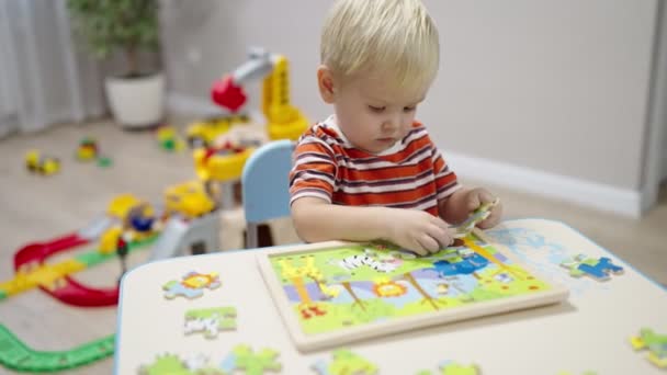 Маленький мальчик с светлыми волосами сидит за столом и собирает деревянные головоломки — стоковое видео