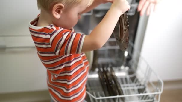 ブロンドの髪の男の子はお母さんが食器洗い機から料理を得るのを助ける座っている — ストック動画