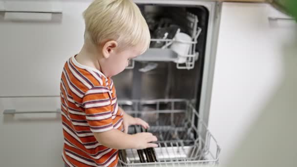 Little boy with blond hair helps mom get the plates out of the dishwasher. — Vídeos de Stock