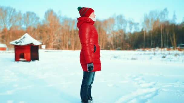 雪に覆われた森の冬の寒さを背景に赤いジャケットの少女が凍った湖に立っています — ストック動画