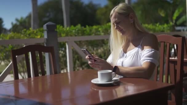 コーヒーを飲みながらカフェに座っている若いブロンドの女性の肖像画 スマートフォンを手に笑顔を浮かべる — ストック動画