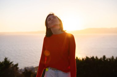 Kırmızı kazaklı genç bir kadın muhteşem bir gün batımında denizin arka planında, romantik bir ruh halinde, mutlu bir şekilde gülümsüyor.