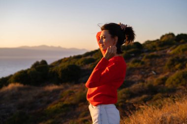 Kırmızı kazaklı genç bir kadın muhteşem bir gün batımında denizin arka planında, romantik bir ruh halinde, mutlu bir şekilde gülümsüyor.