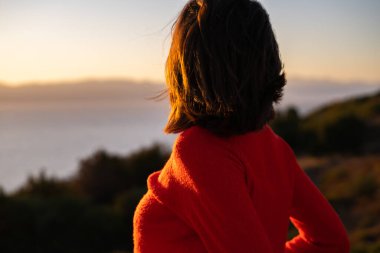 Kırmızı kazaklı genç bir kadın muhteşem bir gün batımında denizin arka planında, romantik bir ruh halinde, mutlu bir şekilde gülümsüyor.