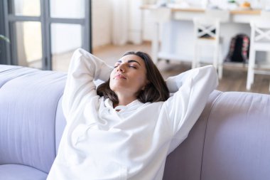 Kanepede beyaz kapüşonlu genç bir kadın oturuyor. Huzur içinde gözleri kapalı, dinleniyor, meditasyon yapıyor.