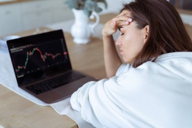 Young woman at home in the kitchen in a white hoodie with a laptop, graph on the screen, upset, unhappy, cryptocurrency falls down clipart