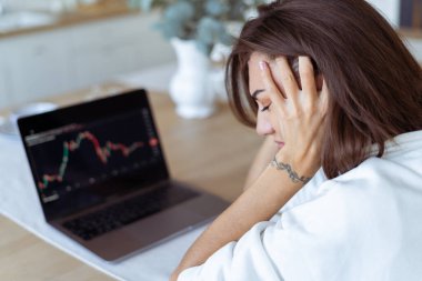 Mutfakta dizüstü bilgisayarı olan beyaz kapüşonlu genç bir kadın, ekranda grafik, üzgün, mutsuz, kripto para birimi düşüyor.