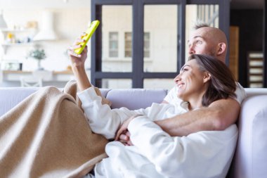 Genç çift evlerinde kanepede rahat bir battaniyenin altında telefonda selfie çekiyorlar.