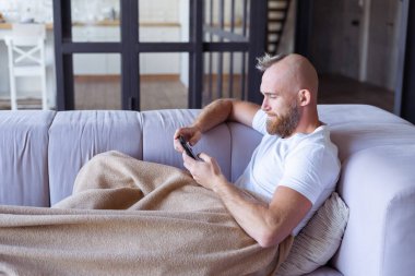 Kanepede oturan genç bir adam sıcak bir battaniyeyle çıkıyor, cep telefonunu tutuyor, haberleri okuyor, video izliyor.