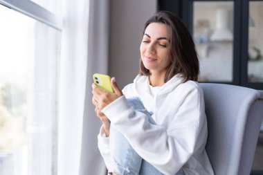 Evinde rahat beyaz kapüşonlu genç bir kadın pencerenin yanındaki koltukta oturuyor, tatmin olmuş, telefon ekranına bakıyor ve gülümsüyor.