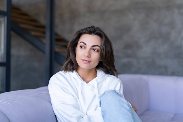 A young woman at home in a white hoodie on the couch sits peacefully, enjoys loneliness, comfort, alone, in a calm mood