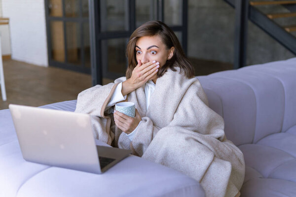 Young woman at home on the sofa in winter under a cozy blanket with a laptop, shocked looks at the screen