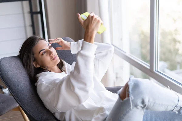 Young woman at home in a cozy white hoodie sits on an armchair by the window, satisfied, looks at the phone screen, takes a selfie