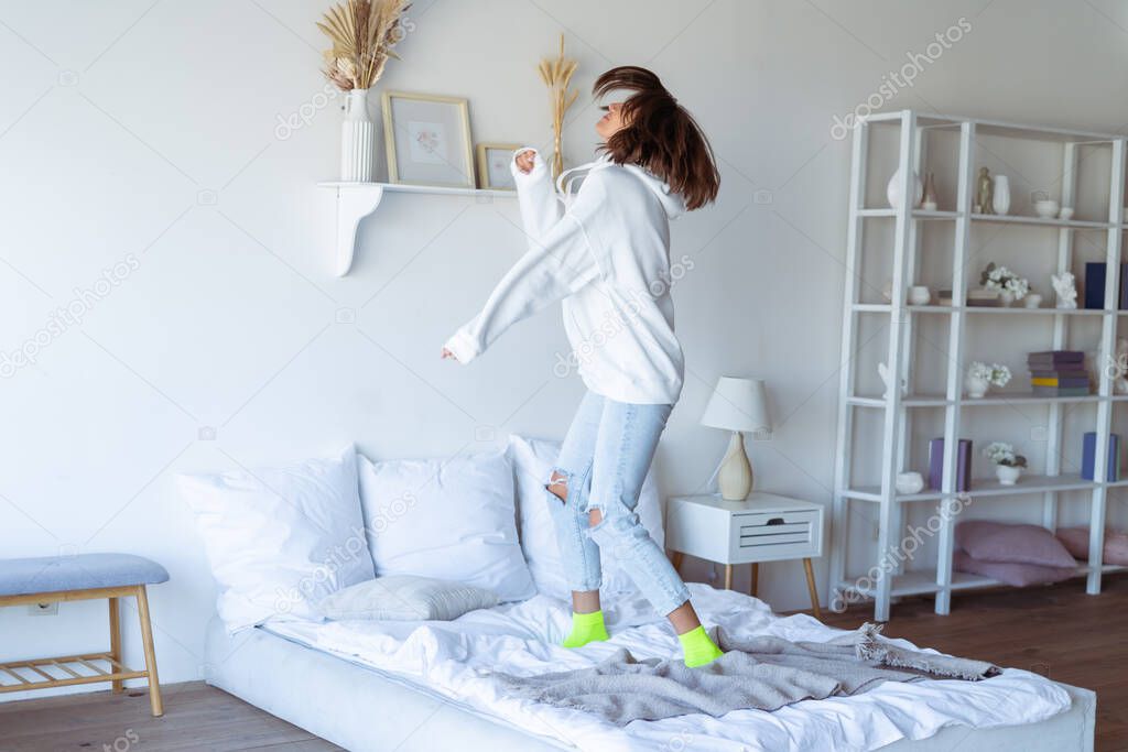 Young woman at home in the bedroom in a warm white hoodie, having fun, jumping on the bed, dancing