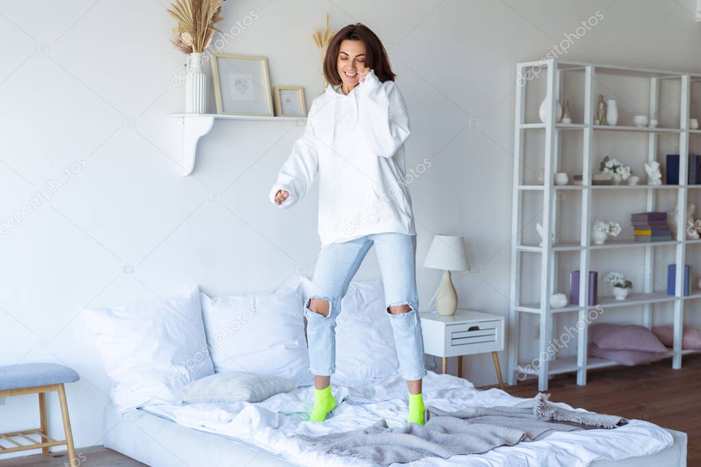 Young woman at home in the bedroom in a warm white hoodie, having fun, jumping on the bed, dancing