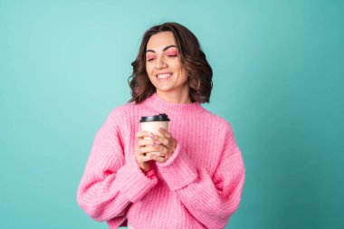 Pembe örgü süveter giyen, turkuaz arka planda parlak makyajlı ve geri dönüşümlü kahve fincanı olan genç bir kadının samimi portresi.