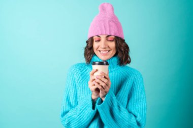 Mavi süveter ve elinde bir fincan sıcak kahve tutan parlak makyajlı pembe şapkalı genç bir kadının samimi portresi.