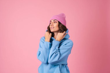 Pembe arka planda, tüylü, kapüşonlu ve pantolonlu, kafasında örülü bir şapka olan genç bir kadın.