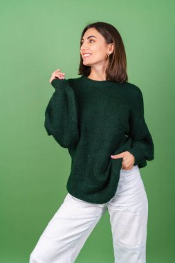 Yeşil arka planda, büyük örülü süveter giyen, tatlı sırıtan genç bir kadın.