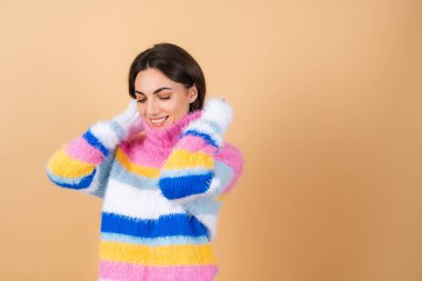 Bej arka planda, parlak renkli, rahat örgü süveteri olan genç bir kadın gülümsüyor.