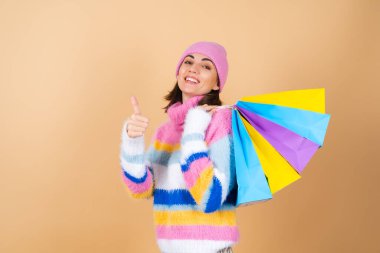 Bej renkli arka planda parlak, çok renkli örgü süveter giyen genç bir kadın. Ekose bir etek, alışveriş paketlerini neşeyle tutuyor.
