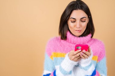 Bej arka planda örgü kazaklı, telefonlu genç bir kadın ekrana bakar ve dudağını ısırır.