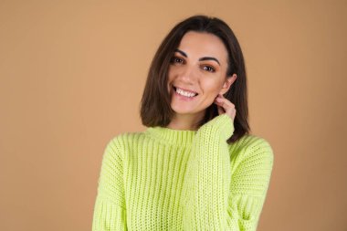 Açık yeşil kazaklı genç kadın bej arka planda mutlu bir şekilde gülümsüyor ve kameraya bakıyor.