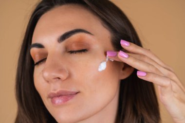 Bej bir arka planda, doğal sıcak makyajlı ve pürüzsüz ciltli genç bir kadının portresi, yüzüne krem sürüyor.