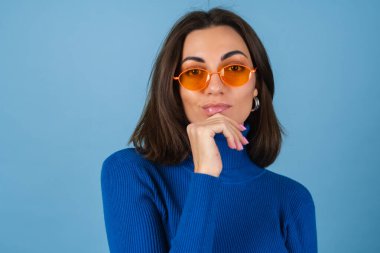 Golf oynayan genç bir kadın mavi arka planda şık turuncu gözlükleriyle iyi bir ruh hali içinde poz veriyor.