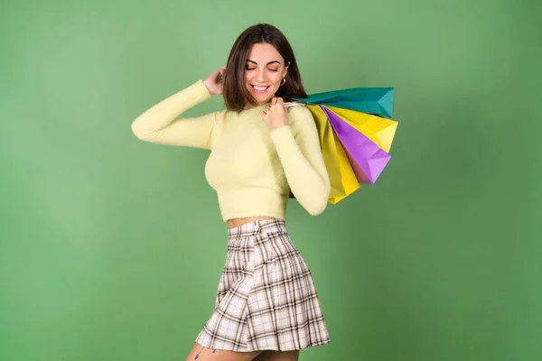 若いです女性で緑の背景に黄色のセーターと遊びスカート保持ショッピングバッグ — ストック写真