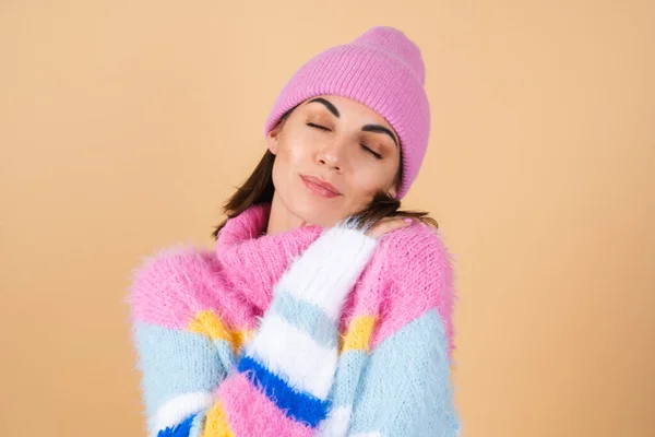 明るいマルチカラーの居心地の良いニットセーターと帽子のベージュの背景に若い女性が快適に身を抱きしめる 暖かさと快適 — ストック写真