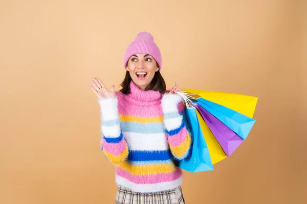 明るい多色の居心地の良いニットセーターのベージュの背景に若い女性は 遊ばれたスカートは興奮して驚きで叫ぶショッピングパッケージを保持しています — ストック写真