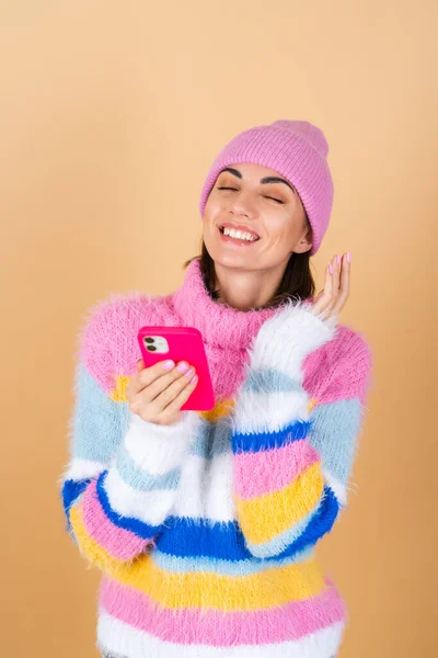 穿着一件针织毛衣 头戴一顶帽子 头戴米黄色背景的年轻女子 戴着一顶带着手机的帽子 带着惊奇和欢乐的笑容 阅读着短信 笑话和祝贺 — 图库照片