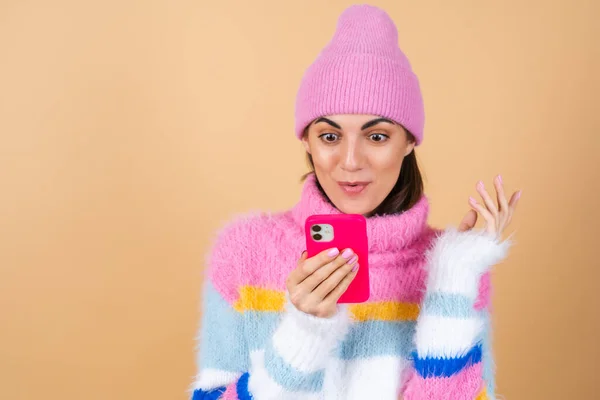 若い女性のベージュの背景にニットセーターと帽子で携帯電話は驚きと楽しさで笑って メッセージ おめでとう — ストック写真