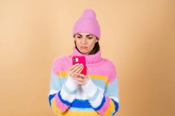 Young woman on a beige background in a knitted sweater and a hat with a mobile phone looks at the screen with disgust displeasure