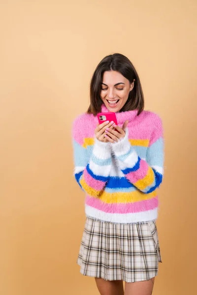 若いです女性上のベージュの背景にニットセーターで携帯電話は 画面を見て笑い陽気に満面の喜び — ストック写真